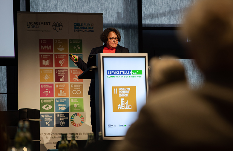 Eine Frau steht an einem Rednerpult; vor ihr unter hinter ihr sind an einer Stellwand die globalen Nachhaltigkeitssymbole zu sehen.